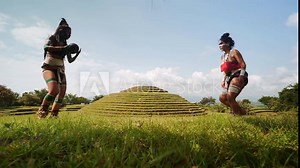 Mayan female warriors in traditional body costumes and face paint engaging in the ancient Mesoamerican ballgame with a dark ball in a grass near ancient ruins in Mexico