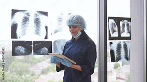 Confident indian woman wearing medical gloves and cap holding digital tablet and standing near window with x-ray results. Professional surgeon reviewing examination results using device indoors.