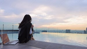 Businesswoman dressed in formal clothes with suit over her shoulder sitting on the edge dangling her feet in swimming pool on skyscraper. Woman using social media on laptop for relax during sunset.