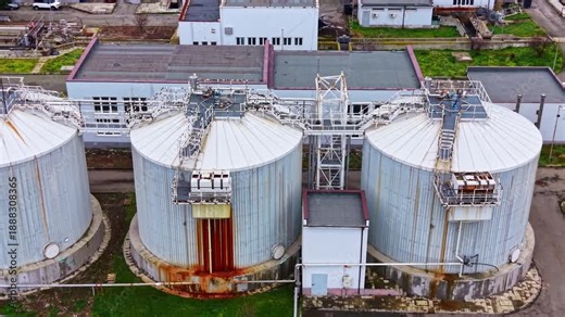 Several large storage tanks stand at an industrial facility. The tanks are metal and have some rust. Surrounding buildings and green areas are visible, showcasing the site layout.