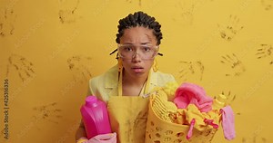 Discontent angry ethnic housewife with dirty face holds laundry basket and detergent looks angrily at camera wears apron big transparent glasses isolated over yellow backgrounf muddy handprints