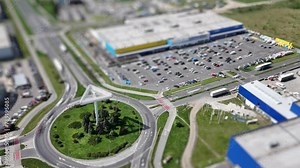 A tilt-shift time-lapse captures cars rapidly moving around a parking lot and road near a prominent advertising pillar with a large shopping center visible in the background.