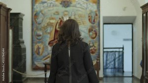 Young woman with curly hair in glasses, plaid dress walks in old library, with blurred tapestry on wall, looks around admiringly. Young specialist, librarian, guide on first working day at new job.