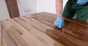 Male carpenter in rubber gloves polishes with varnish wooden table surface at workplace in apartment office. Man uses liquid lacquer slow motion