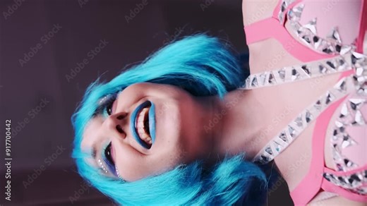 Beautiful girl with blue hair and blue lips licks a bright heart-shaped lollipop. View from below. Girl in a pink bra decorated with rhinestones, a short white skirt and white gloves. Vertical video