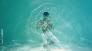 Underwater shot of young man entangled in chains. Man screams and tries to get out of blue water column with glare. Emotions of suffering, captivity of chains. Subaquatic shot, close up in slow motion