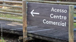 Wooden sign with arrow pointing left to shopping center access, placed on a small wooden footbridge in a park