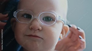 One year old infant with strabismus and glasses looks at camera and smiles. Caucasian baby has squint, bad vision. White child with poor eyesight. Closeup, portrait of kid girl with an eye disease