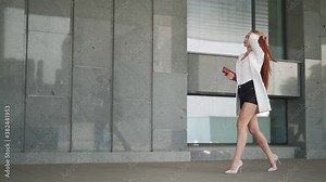 Beautiful confident business woman with red hair in fashion clothes walk to work near office. Young lady enjoying successful corporate career. Elegant female entrepreneur walks downtown. Slow motion