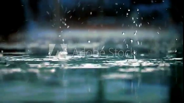 Close-up slow-motion footage of raindrops striking a shallow puddle, forming elegant crowns, bubbles, and ripples on the water surface. Blurred green foliage in the background.
