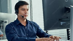Young competent asian male call center agent working at his computer while simultaneously speaking with customers. Concept of an operator, customer service agent working in the office with headset.