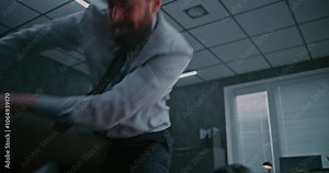 Aggressive businessman loses his temper in a fit of rage. Furious male office employee smashes computer monitor with keyboard, throws things off the desktop and yells. Financial crisis or dismissal.