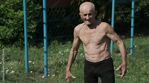 Strong very old man with bare torso demonstrates arms muscles on athletic ground. Thin old man strains muscles of arms showing biceps and belly press