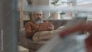 Medium long shot of older Caucasian man with long beard lounging on sofa in office, using tablet computer, finger of unrecognizable person bending down parts of venetian blinds, peeping through glass