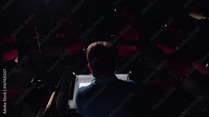 Mid aged conductor alone in dark orchestra pit studies sheet music and tweets the tune. Indoor. Medium. Camera from behind, and from above.
