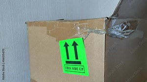 Male hand sticking green "this side up" sticker with arrow symbol on delivery cardboard box before shipping. Handle with care concept