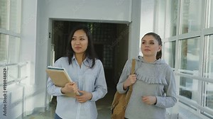 Two young beautiful female students walking in wide lighty corridor of college discussing homework smiling positively. Asian girl is holding books and papers.