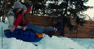 Two attractive happy young women smile sledging on snow slope towards sunny house. Winter holiday leisure slow motion.