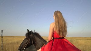 Girl horseback rider wearing long red dress riding horse on country road in the evening. Bright gown blowing in the wind. Rear view of horsewoman on black stallion walking across dry grass field.
