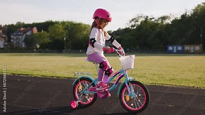 A beautiful, sweet girl in a pink helmet and pink knee and elbow pads is learning to ride a bicycle at the stadium. The girl is delighted with the new experience on a sunny summer day.