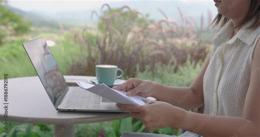 Asian adult woman multitasking by reviewing printed report and working on laptop at natural outdoor cafe, balancing professional career with freedom in remote digital lifestyle and scenic nature
