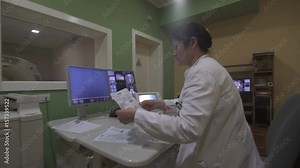 Female radiology technician and radiologist are working at computers at scanner control room, concept scanning the patient and review of x-ray, steady cam shot, indoors, real scene.