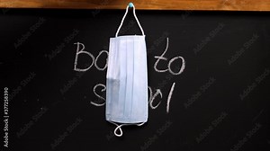 white chalk lettering on black chalkboard back to school, hand hangs a disposable medical mask on the chalk board, concept of self-isolation during an epidemic, distance learning