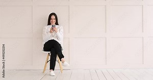 Happy woman text messaging using mobile phone sitting on chair against wall