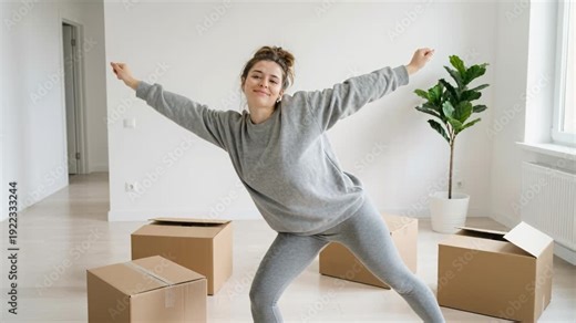Woman stretches and celebrates in a new empty apartment with moving boxes, showing happy relocation and moving day