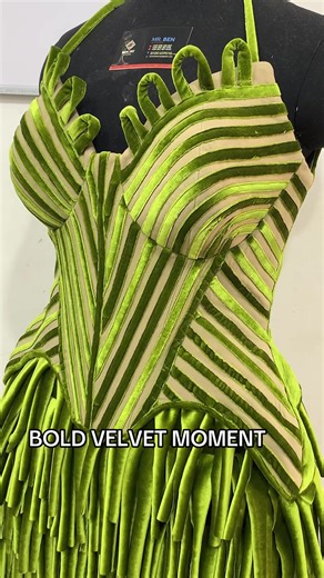 A structured avant-garde mini dress crafted in rich olive green velvet. The bodice features a sculpted corset silhouette with diagonal striped panel detailing that enhances the waist and creates a striking visual contour. The sweetheart neckline is elevated with dimensional looped velvet appliqués, adding a dramatic architectural touch. The peplum waist extends into a textured mini skirt adorned with layered velvet tassel fringe, creating movement and depth. The combination of structure and flui