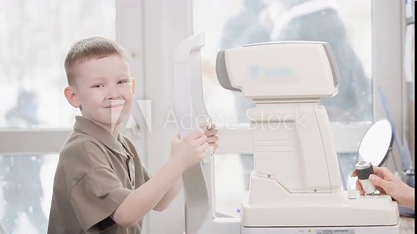 Tracking shot of young boy smiling during eye exam with autorefractor in bright optometry clinic by window.