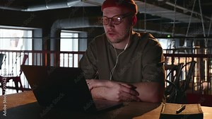 A male remote worker, communicates with his management or a customer by video call using a laptop and white wired headphones from a dark cafe room. Freelance millennial hipster with glasses. 4k
