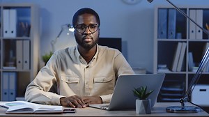 Free stock video - Young office worker sitting at the office desk looking and smiling at camera in the office at night