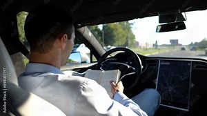 Male businessperson reading book during riding on electrical vehicle with autopilot at urban road. Successful businessman improving his knowledge while riding an autonomous self driving electric car