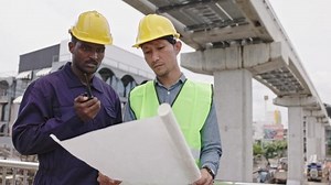 Structural engineers and construction workers are holding blueprints inspecting structure. sky train track construction in city. Blue collar wears a safety hardhat while working on construction site.