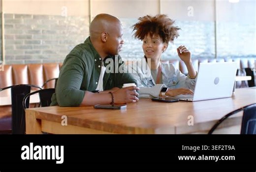 African couple sitting at cafe table, reviewing laptop screen, discussing work, man holding coffee. Collaboration, conversation, device, smartwatch, coffeehouse, wooden, bench Stock Video Footage - Alamy