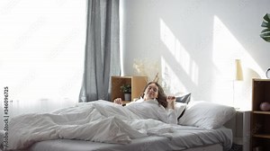 Tracking shot of beautiful brunette woman waking up in her bed at home, sitting up and smiling while enjoying bright morning sunlight shining through the window Stock Video
