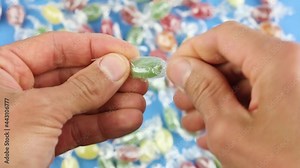 hands unfold one green candy lollipop from a transparent wrapper, on background of many colored candies, unhealthy sugar sweets, sugar addiction