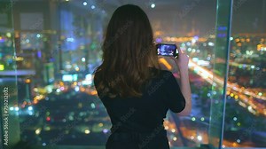 Woman make panoramic shot of night big city lights. Female standing on top floor viewpoint, holding camera photograph the urban skyline. The cityscape features tall buildings, cars, and streets roads