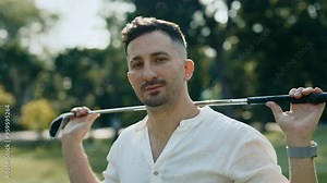 Portrait of attractive golf player with beard looking at camera in sunny park. Cheerful golfer holds steel club behind neck standing on course. Athlete enjoys training outdoors