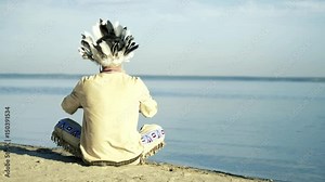 An Indian in a national costume plays a flute on the river bank 4k