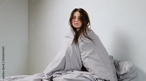 Sick young woman sitting up in bed, wrapped tightly in a gray blanket, looking pale and tired, with messy hair and a weak expression, recovering from illness in a minimal room.