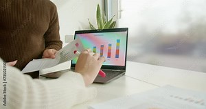 Sitting at a desk next to the window, two businesspeople work together, analyzing reports and graph charts, aiming for a understanding of profit and sales statistics in the corporate environment.