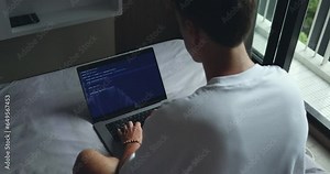 Digital Workspace. Man Typing Programmer Code on Laptop, Seated on Bed, Rear View. IT Freelancer writing code on his laptop from the comfort of his bed