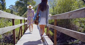 Bridge, vacation and friends walking in forest in summer, travel or back outdoor. Rear view, holiday or women in woods by path with hat on adventure, hiking or explore in nature together in Australia