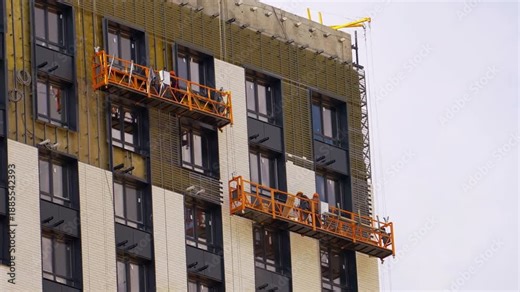 Construction workers installing modern facade panels on high-rise residential building using suspended platforms, exterior insulation and cladding system, urban real estate development concept