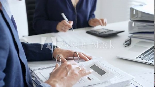 Two professional accountants collaborating in a modern office, calculating taxes and analyzing financial data with calculators, documents, and a laptop. Audit and taxes in business