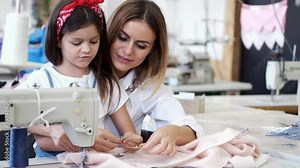 The woman helps the girl. Little girl makes clothes. They use sewing machine.