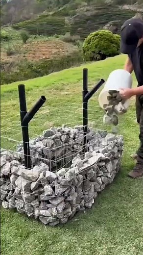 pouring the grey stones into a wire gabion basket to build a natural garden wall in the green field