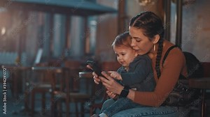 Don't bother me please. Busy young mum sitting at the street cafe with her cute little kid and using smartphone, trying to find address, text, open social network or websites. Child very interested